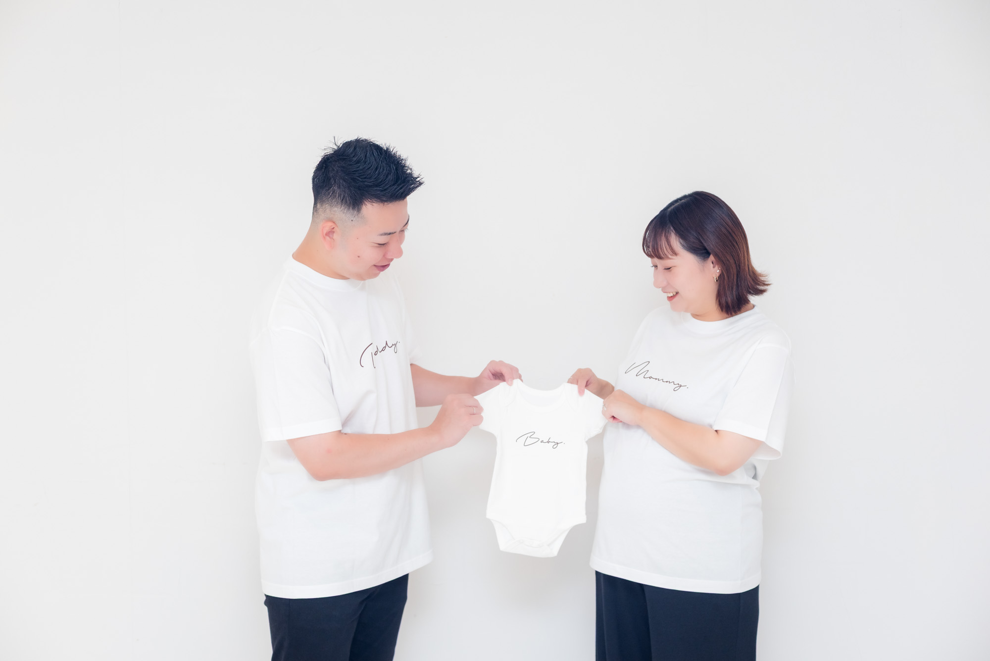 白いTシャツ姿のパパとママが、Babyと書かれた小さなベビー服を笑顔で広げている写真