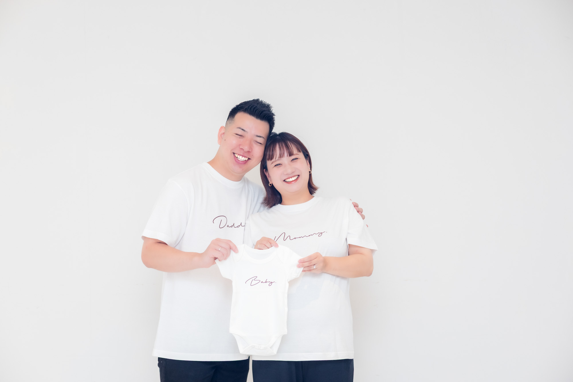 白いTシャツ姿のパパとママが、小さなベビー服を持って寄り添っている写真
